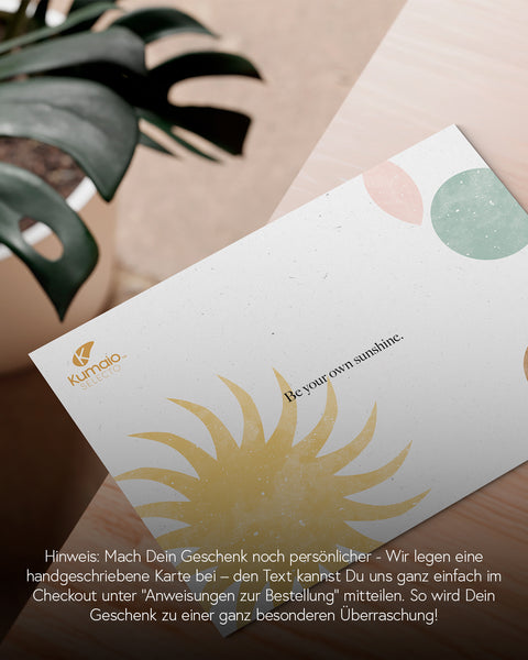 Alt-Text: Eine handgeschriebene Karte mit einem Sonnensymbol und dem Spruch „Be your own sunshine“ liegt auf einem Tisch. Daneben eine grüne Pflanze im Hintergrund. Text im Bild erklärt, dass Geschenke durch eine personalisierte Nachricht auf der Karte ergänzt werden können.