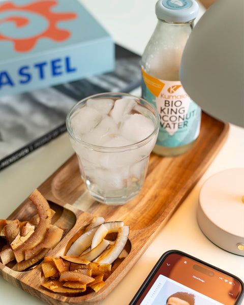 Holztablett mit Kokoschips, einem Glas mit Eis und einer Flasche Kumaio Bio King Coconut Water, daneben ein Smartphone mit geöffneter Musik-App.