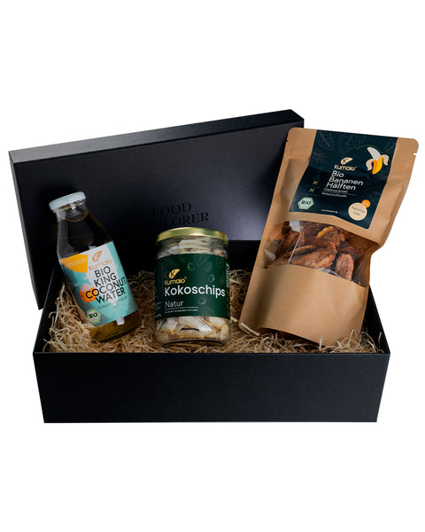 Schwarze Geschenkbox mit drei Bio-Produkten: eine Flasche King Coconut Water, ein Glas Kokoschips Natur und eine Tüte getrocknete Bio-Bananenhälften. Die Produkte sind dekorativ auf Holzwolle gebettet.