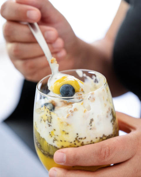 Hand hält ein Glas mit Chia-Pudding, Sweet Jackfruit, Joghurt und Blaubeeren, ein Löffel wird gerade zum Mund geführt.