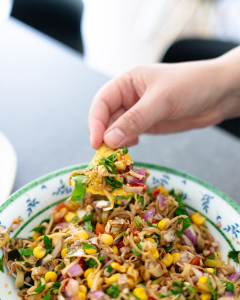 Hand hält Tortilla-Chip mit bunter Ceviche aus Bananenblüten, Mais, Kräutern und Zwiebeln über einer Schüssel.
