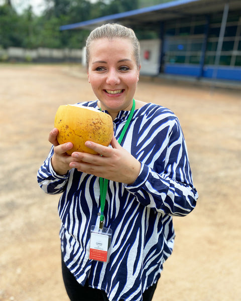 Eine Frau mit blauer und weißer Zebra-Bluse hält eine geöffnete gelbe Kokosnuss und lächelt in die Kamera.