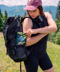 Kumaio® Bio Kokoschips Ananas im 110-g-Beutel als Snack für unterwegs, verstaut am Wanderrucksack vor alpiner Berglandschaft.