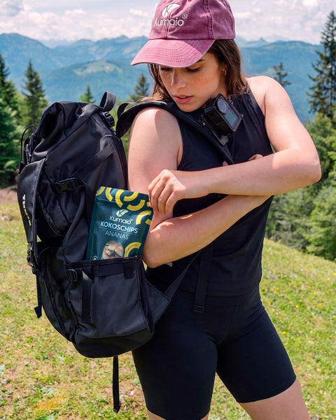 Kumaio® Bio Kokoschips Ananas im 110-g-Beutel als Snack für unterwegs, verstaut am Wanderrucksack vor alpiner Berglandschaft.