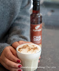 Kumaio Chai Sirup und frisch zubereiteter Chai Latte mit Milchschaum und Zimt, gehalten von einer Person mit grauem Pullover.