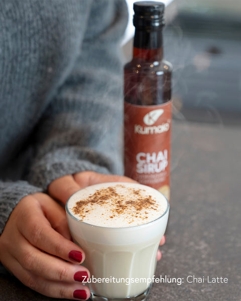 Kumaio Chai Sirup und frisch zubereiteter Chai Latte mit Milchschaum und Zimt, gehalten von einer Person mit grauem Pullover.