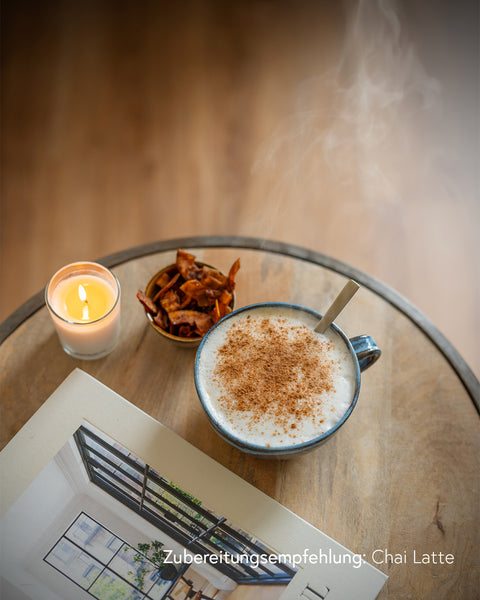 Tasse heißer Chai Latte mit Zimt bestreut auf Holztisch, daneben Kerze, Snackschale und aufgeschlagenes Magazin, Zubereitungsempfehlung mit Kumaio Bio Chai Sirup.