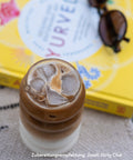 Glas mit Iced Dirty Chai auf Eiswürfeln, zubereitet mit Kumaio Bio Chai Sirup, daneben gelbes Ayurveda-Buch und Sonnenbrille.