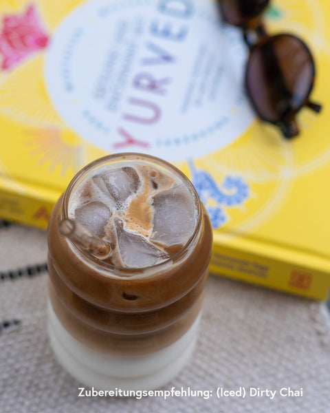 Glas mit Iced Dirty Chai auf Eiswürfeln, zubereitet mit Kumaio Bio Chai Sirup, daneben gelbes Ayurveda-Buch und Sonnenbrille.