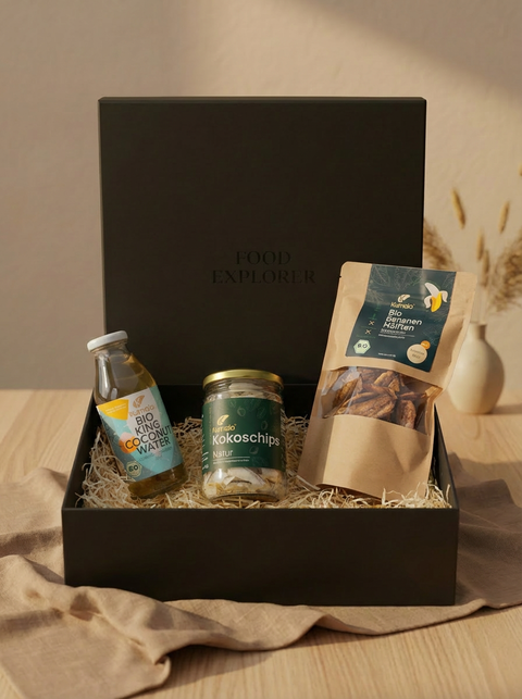 Geschenkbox mit Kumaio Bio King Coconut Water, Bio Kokoschips Natur und Bio Bananen Natur, dekorativ präsentiert in schwarzer Box mit Holzwolle auf hellem Holztisch.