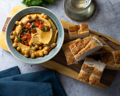 Hummus, garniert mit ganzen Kichererbsen, frischen Tomatenwürfeln und Kräutern. Dazu ein Schuss Kumaio Kokos Aminos Ingwer-Zitronen Gewürzsauce, serviert mit frischem Brot auf einem Holzbrett.