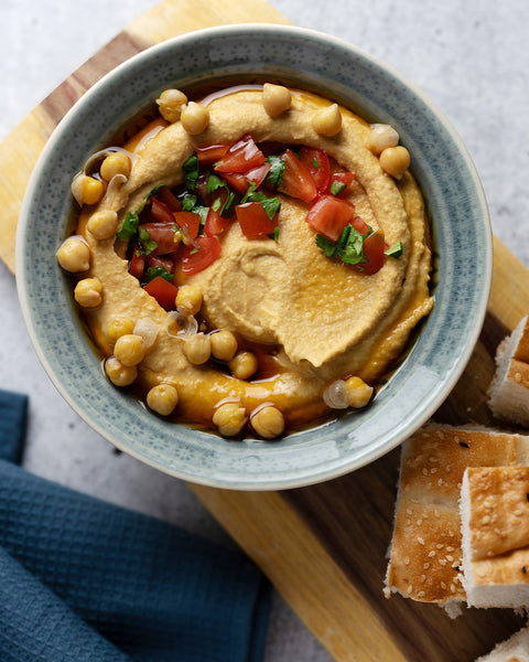Hummus, garniert mit ganzen Kichererbsen, frischen Tomatenwürfeln und Kräutern. Dazu ein Schuss Kumaio Kokos Aminos Ingwer-Zitronen Gewürzsauce, serviert mit frischem Brot auf einem Holzbrett.
