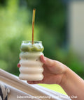 Hand hält ein Glas mit zweischichtigem Iced Matcha Latte mit Strohhalm, unten Milch, oben grüner Matcha, vor unscharfem Naturhintergrund im Sommerlicht.