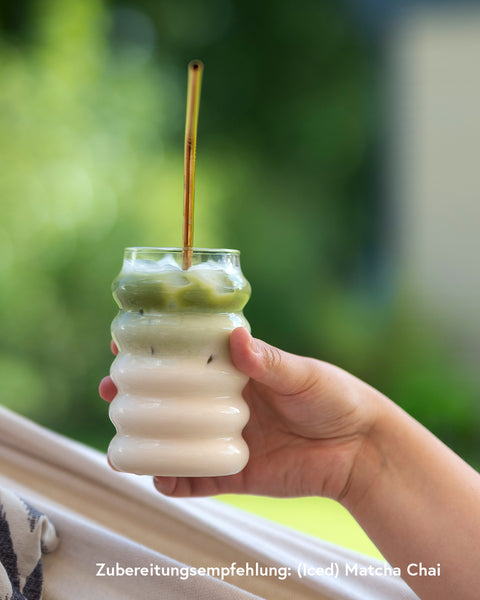 Hand hält ein Glas mit zweischichtigem Iced Matcha Latte mit Strohhalm, unten Milch, oben grüner Matcha, vor unscharfem Naturhintergrund im Sommerlicht.