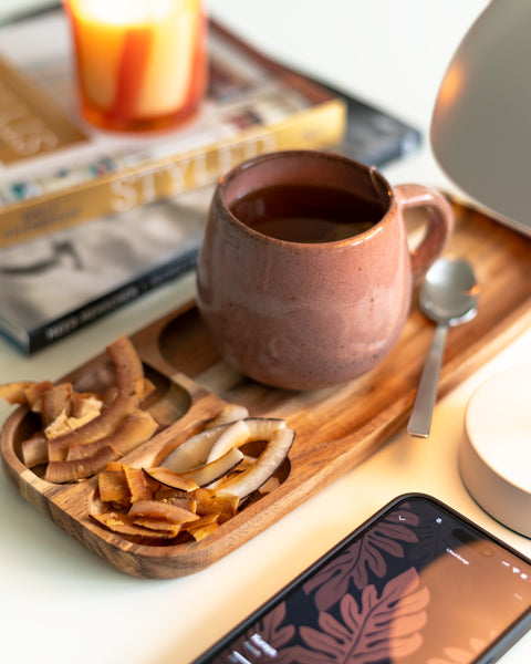 Holztablett mit einer Tasse Tee und verschiedenen Sorten Kokoschips, umgeben von einem Smartphone und Zeitschriften. Perfektes Arrangement für eine gemütliche Snack-Pause, ideal für Wellness- und Genussmomente.