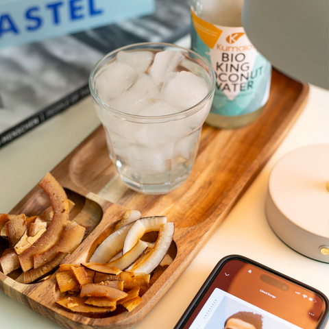 Holztablett mit verschiedenen Sorten getrockneter Kokoschips, einem Glas mit Eiswürfeln und einer Flasche Kumaio Bio King Kokosnuss Wasser, daneben ein Smartphone mit geöffneter Musik-App. Erfrischendes und gesundes Setup für eine Auszeit oder Snack-Pause.