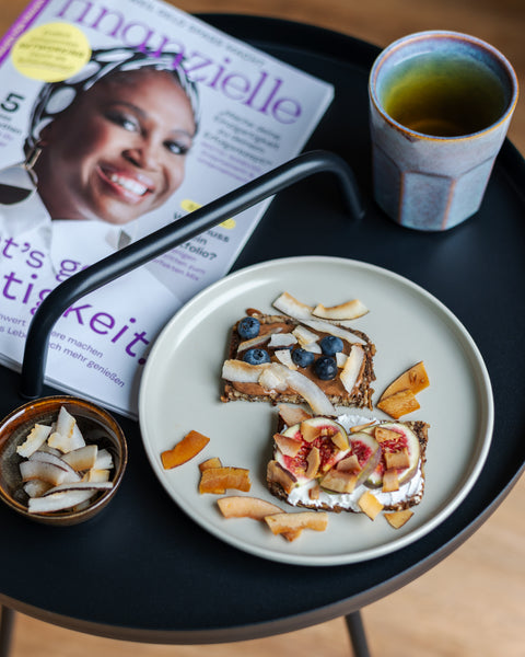 Frühstücksteller mit Vollkornbrot, belegt mit Feigen, Blaubeeren und knusprigen Kokoschips, neben einer Tasse Tee und einer Zeitschrift auf einem schwarzen Tisch. Gesundes und leckeres Frühstück für den perfekten Start in den Tag