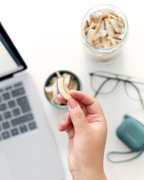 Nahaufnahme einer Hand, die eine knusprige Kokoschips-Scheibe hält, mit einem offenen Laptop und einem Glas voller Kokoschips im Hintergrund. Perfekter Snack für unterwegs oder im Büro, ideal für gesundheitsbewusste Genießer.