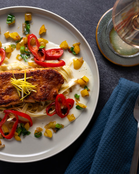 Ein Teller mit gebratenem Lachs auf Nudeln, garniert mit roten Paprikascheiben, gelben Paprikawürfeln und fein geschnittenem Ingwer.