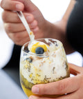 Hand hält ein Glas mit Chia-Pudding, Sweet Jackfruit, Joghurt und Blaubeeren, ein Löffel wird gerade zum Mund geführt.