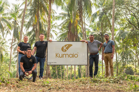 Kumaio Team in Sri Lanka steht vor einem Firmenschild auf einer Kokosplantage in Dompe, umgeben von Palmen – nachhaltige Zusammenarbeit und Ursprung der Bio-Produkte.