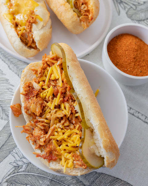 Hotdog-Brötchen mit BBQ-Jackfruit, Gewürzgurke und geriebenem Käse auf weißem Teller. Im Hintergrund weitere belegte Brötchen.
