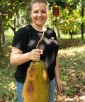 Eine Frau in schwarzem Kumaio-T-Shirt hält eine große Jackfrucht in der Hand, während sie in einem Obstgarten lächelt.