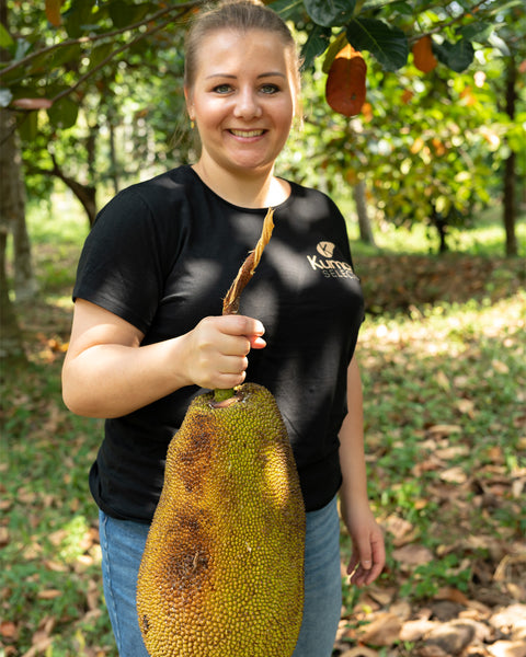 Eine Frau in schwarzem Kumaio-T-Shirt hält eine große Jackfrucht in der Hand, während sie in einem Obstgarten lächelt.